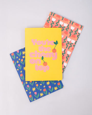 set of three notebook set: dark blue with tulip print, yellow with 'you're the cherry on top' typography, and pink mid century tulips