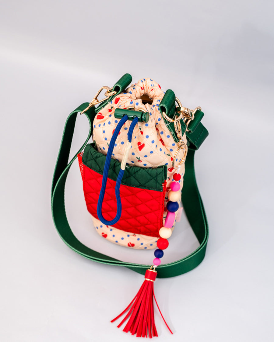 cinched cream quilted water bottle sling with red and green front pouch, green adjustable strap and colorful beaded tassel keychain