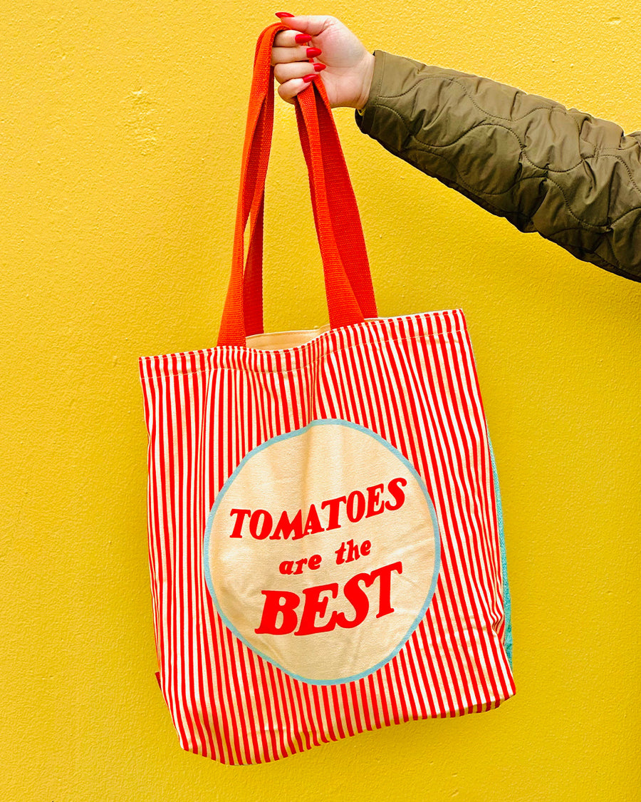 model holding reverse side of tote bag that says 'tomatoes are the best' with yellow and red vertical stripes and red handle