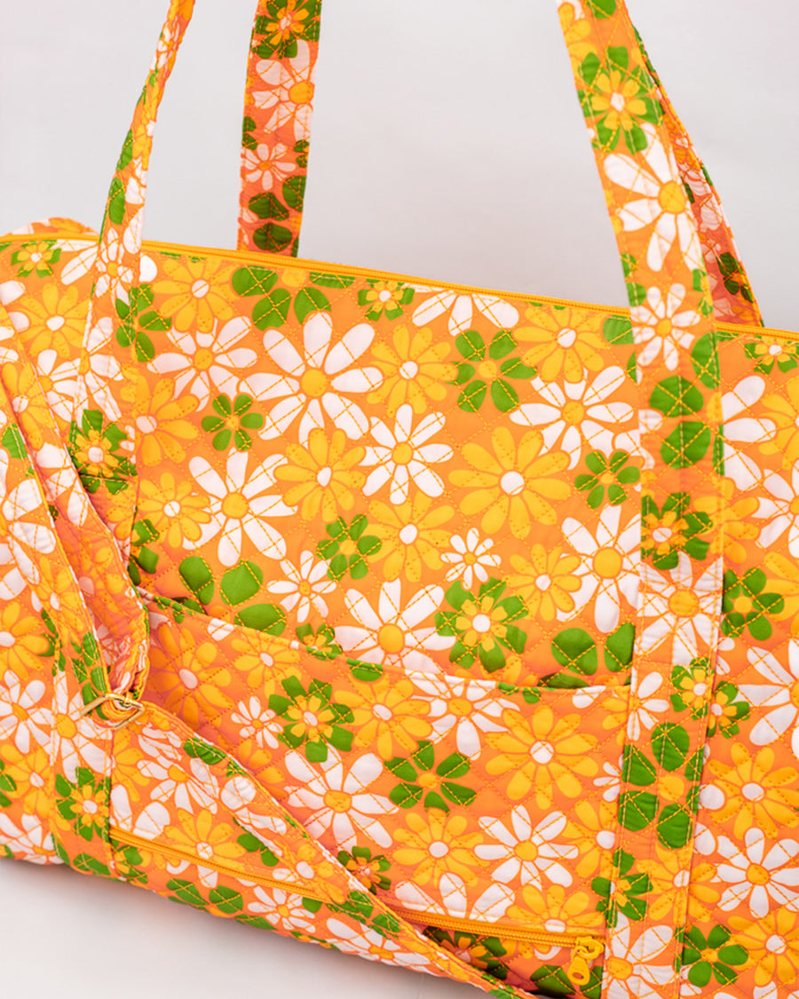 up close of orange quilted duffel bag with handles and adjustable strap and has an all over yellow, white, and green retro floral print