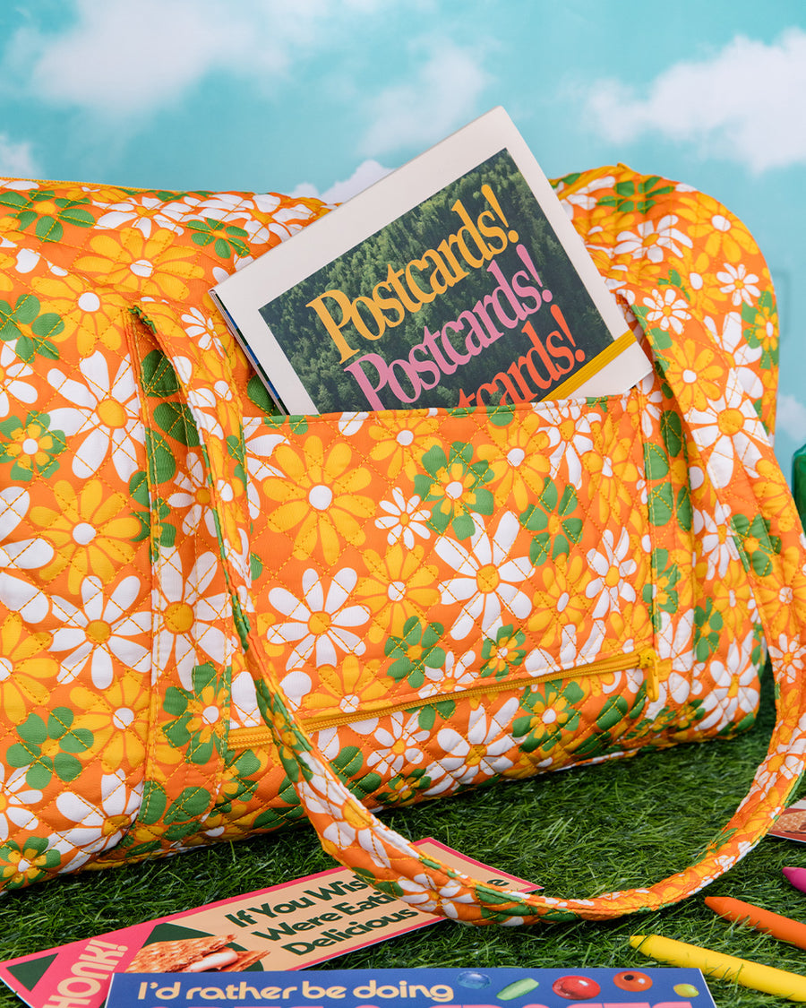 orange quilted duffel bag with handles and adjustable strap and has an all over yellow, white, and green retro floral print with postcard book inside front pocket