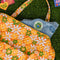 orange quilted duffel bag with handles and adjustable strap and has an all over yellow, white, and green retro floral print with jean jacket with patches sticking out of it