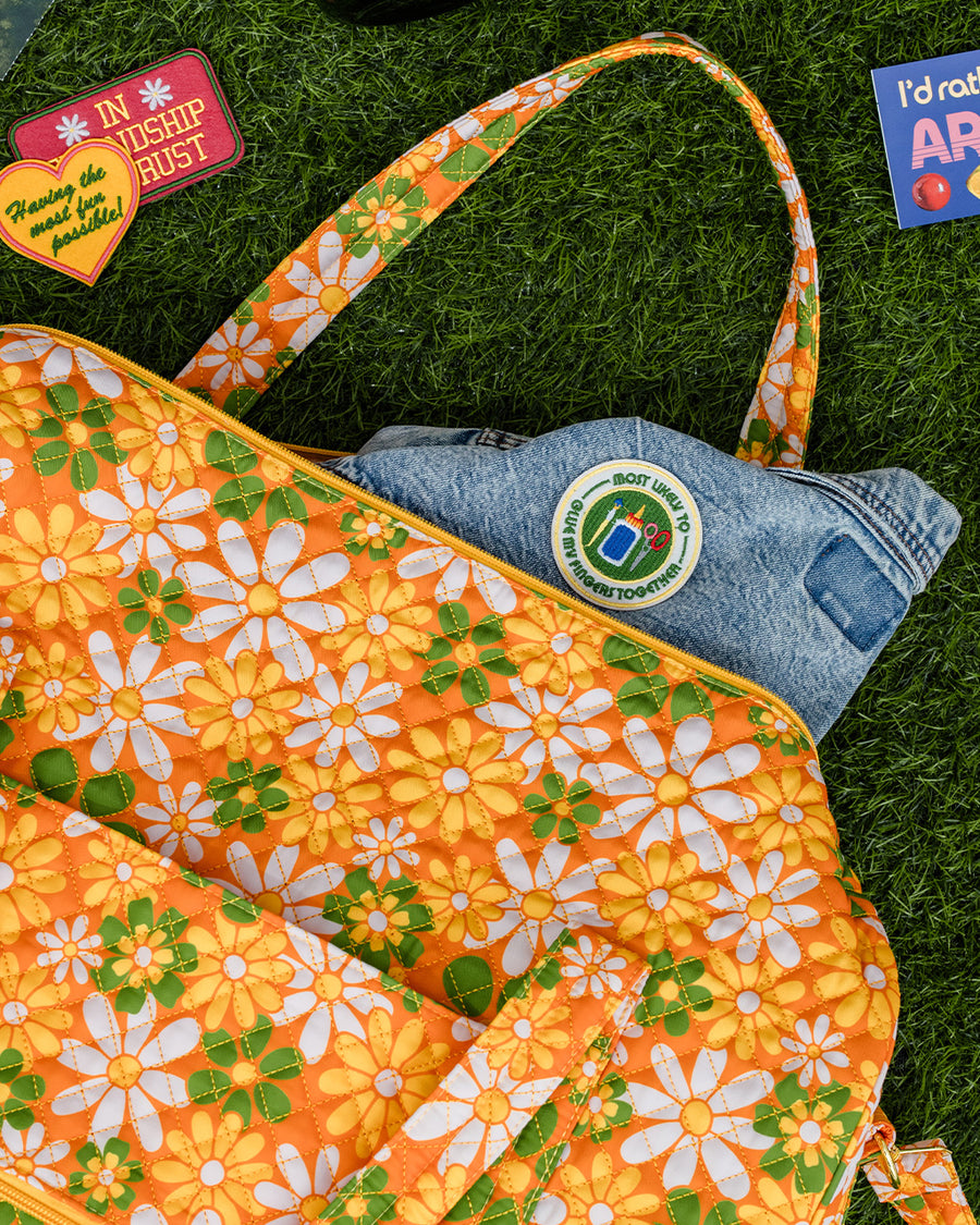 orange quilted duffel bag with handles and adjustable strap and has an all over yellow, white, and green retro floral print with jean jacket with patches sticking out of it