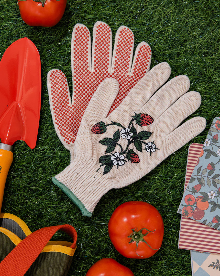 cream gardening gloves with white flower and strawberry print and green trim in the grass surrounded by gardening tools