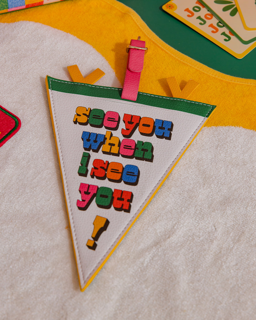 cream pennant luggage tag with colorful 'see you when i see you!' front and pink, yellow and green trim on a towel