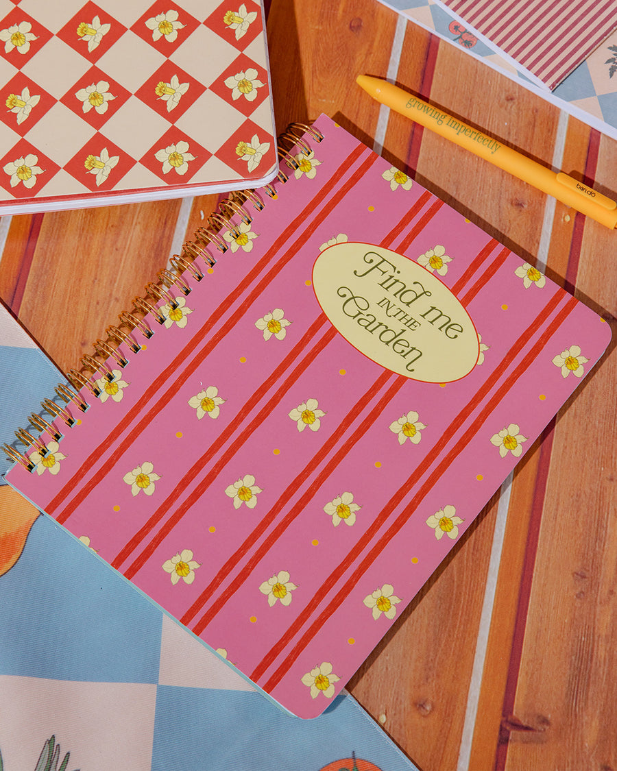 pink mini notebook with vertical double red stripes, realistic daffodil print and cream circle that says 'find me in the garden' on a table