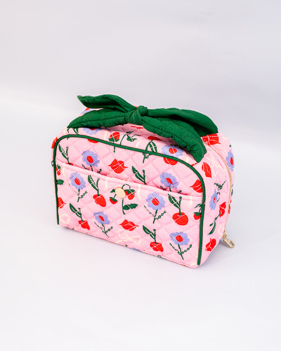 side view of pink quilted toiletry bag with green bow handle and all over periwinkle flower, cherries, bow and lip print