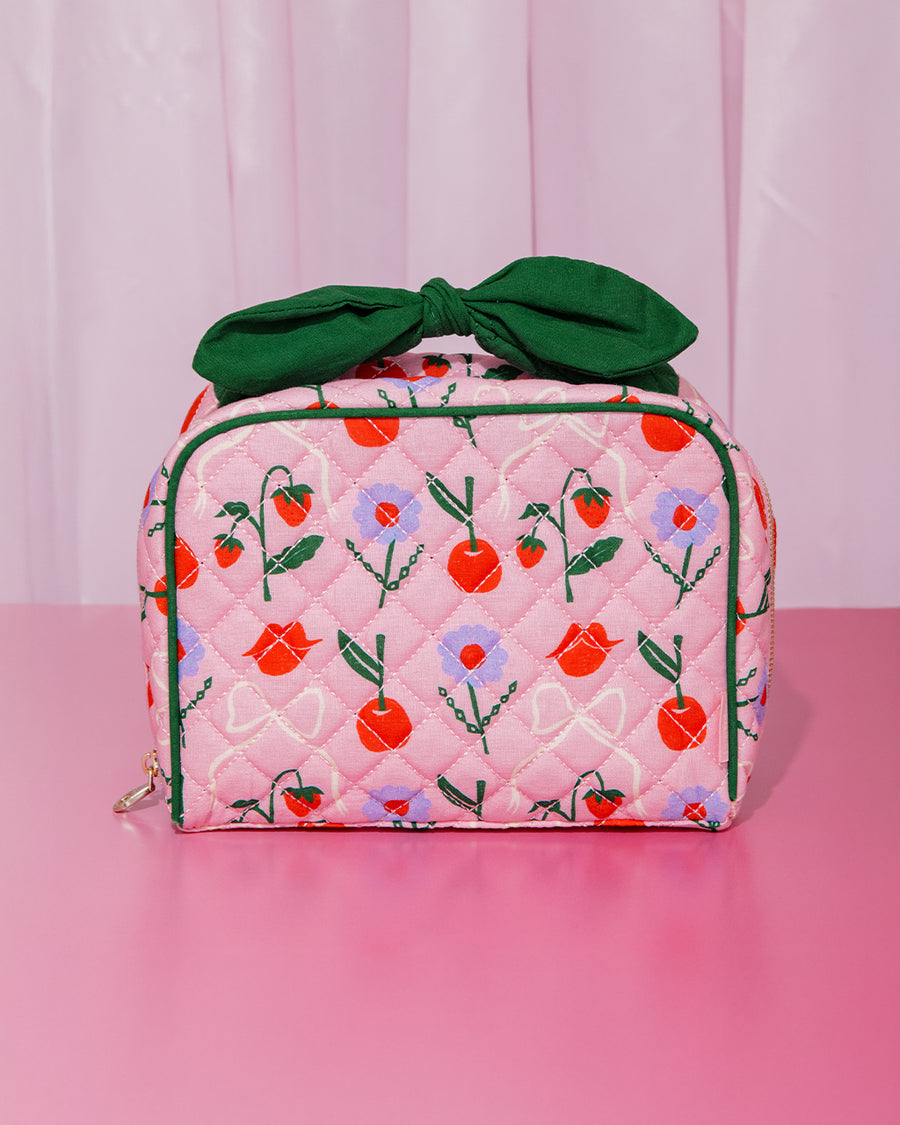 pink quilted toiletry bag with green bow handle and all over periwinkle flower, cherries, bow and lip print on a pink counter and light pink backdrop