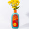 teal ceramic vase with red and yellow 'sunblock 30 SPF' label and yellow flowers inside