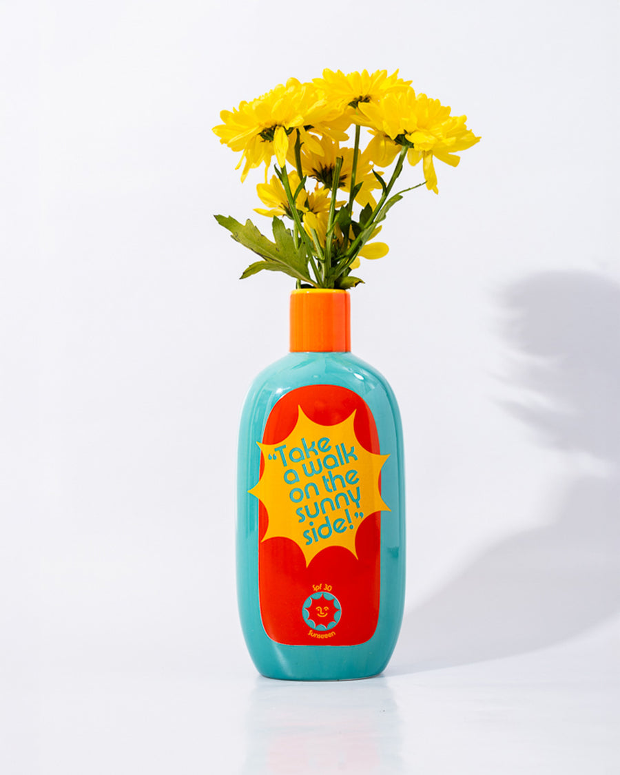 'take a walk on the sunny side' back view of teal ceramic vase with red and yellow 'sunblock 30 SPF' label and yellow flowers inside