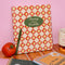 cream waterproof notebook with red diamond and daffodil print with green 'planting seeds of possibility' on a pink wall