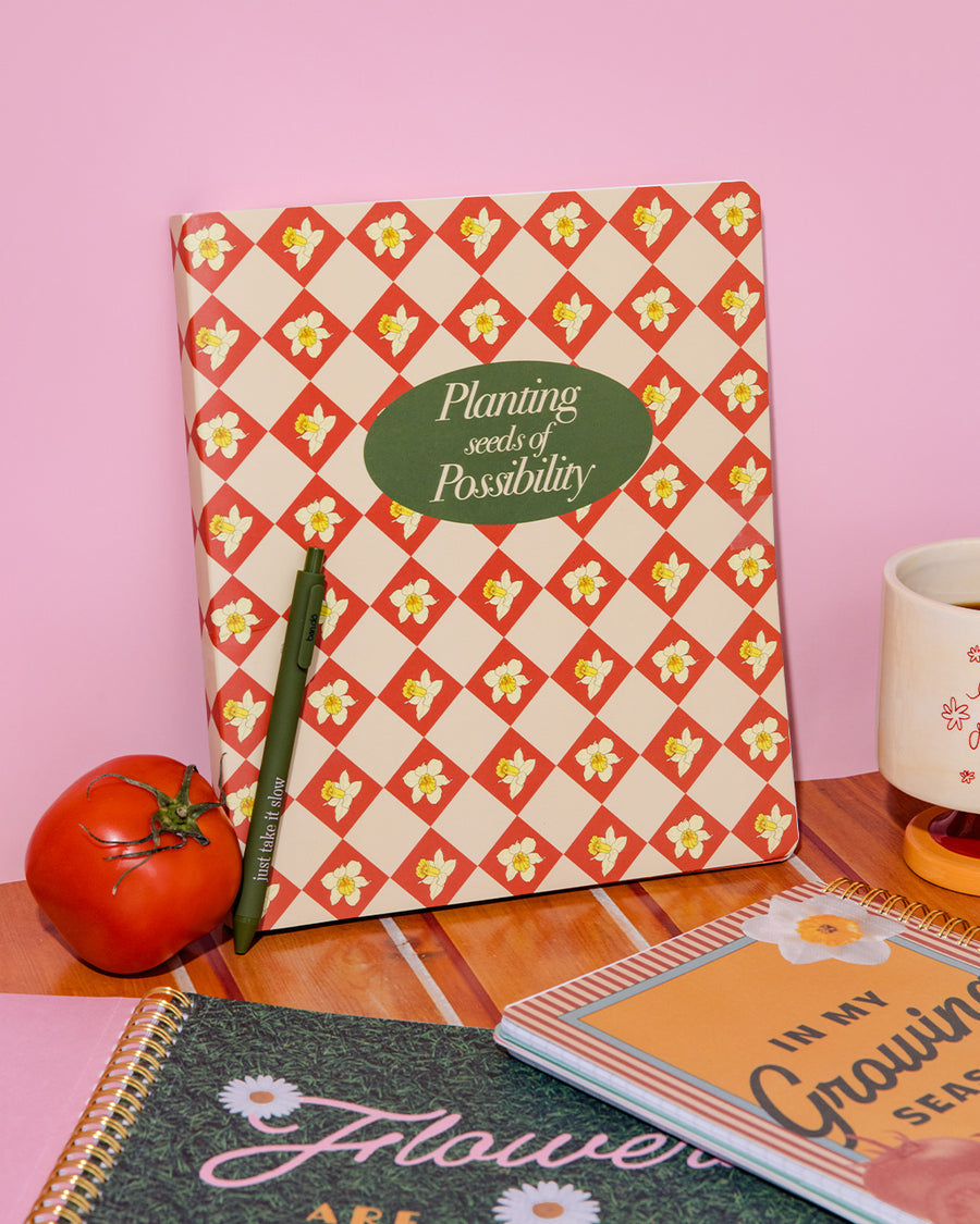 cream waterproof notebook with red diamond and daffodil print with green 'planting seeds of possibility' on a pink wall