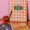 cream waterproof notebook with red diamond and daffodil print with green 'planting seeds of possibility' on a pink wall