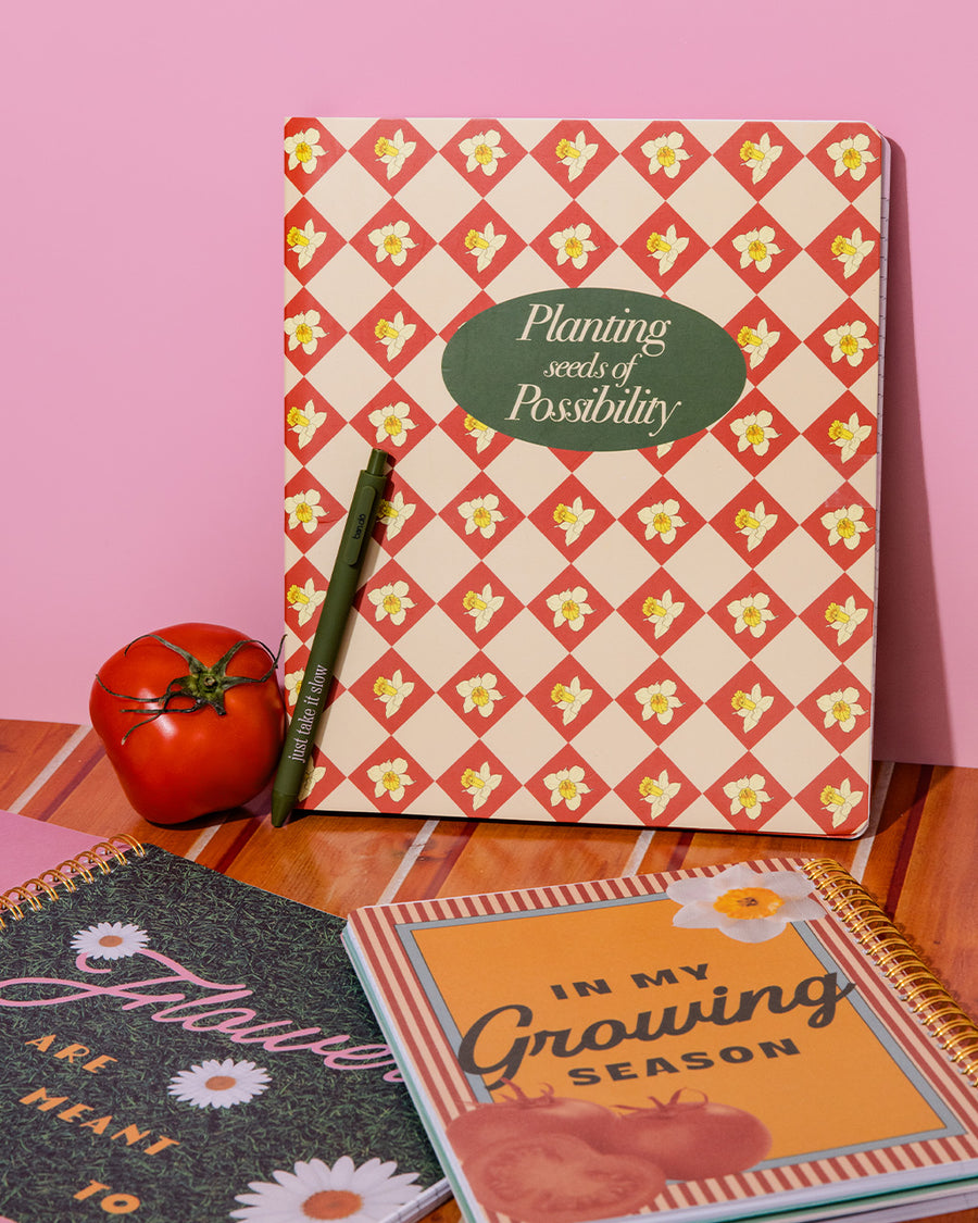 cream waterproof notebook with red diamond and daffodil print with green 'planting seeds of possibility' on a pink wall