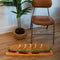 long hoagie shaped throw rug with lettuce, cheese and 'deli meat' center laying on a floor with a chair. and plant behind it