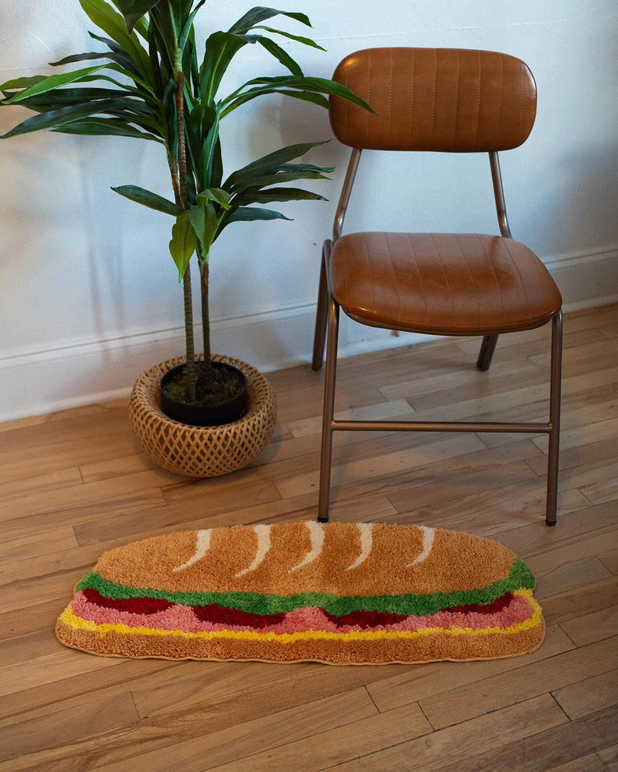 long hoagie shaped throw rug with lettuce, cheese and 'deli meat' center laying on a floor with a chair. and plant behind it