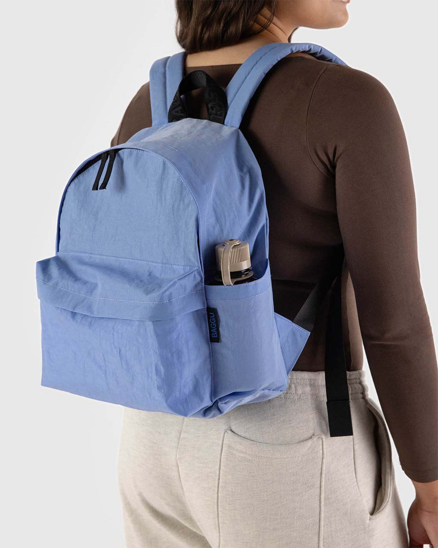 model wearing cornflower medium nylon backpack with large zipper compartment, front zipper pocket, and side drink holder