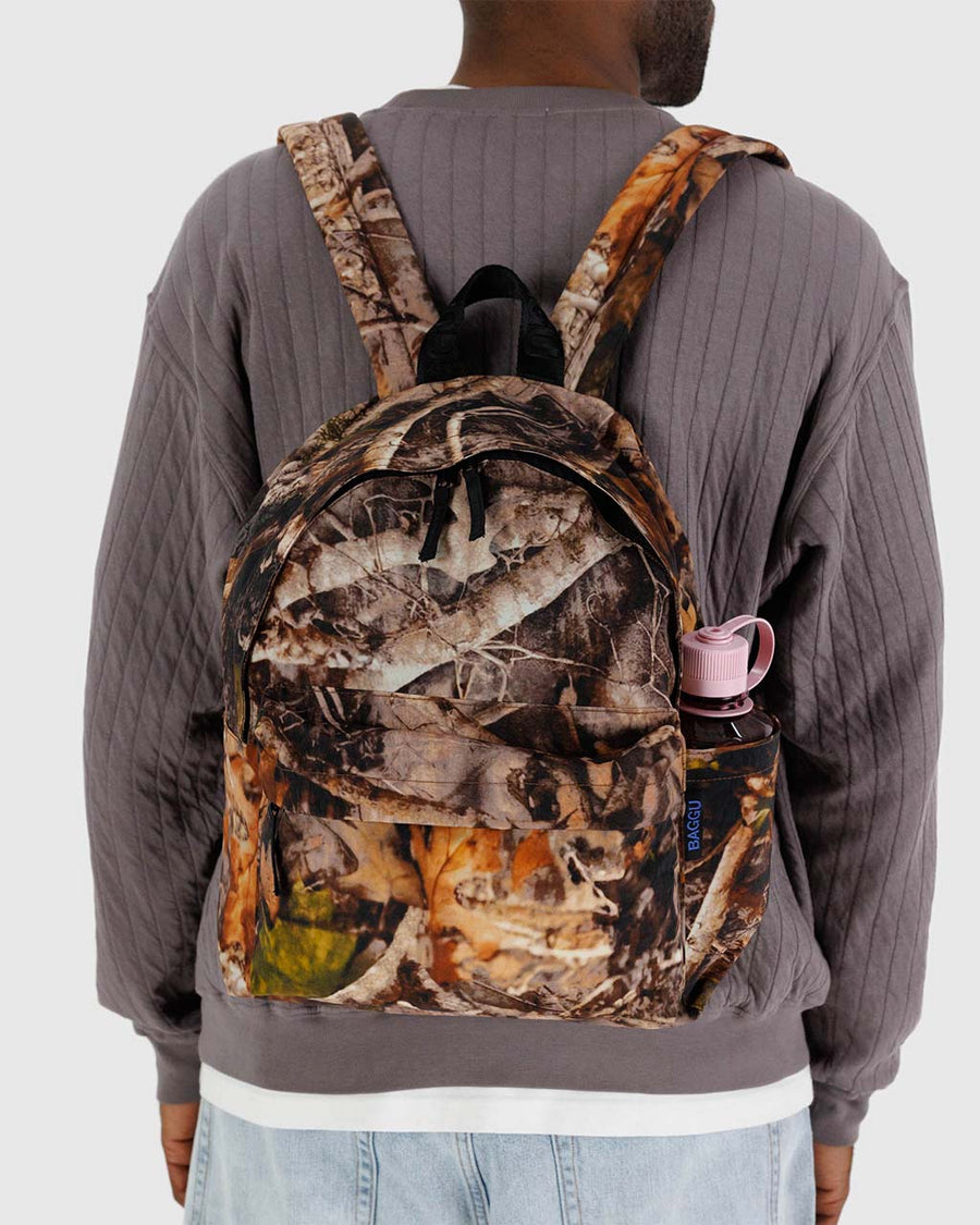 model carrying realistic forest print nylon backpack with large zipper compartment, front zipper pocket, and side drink holder