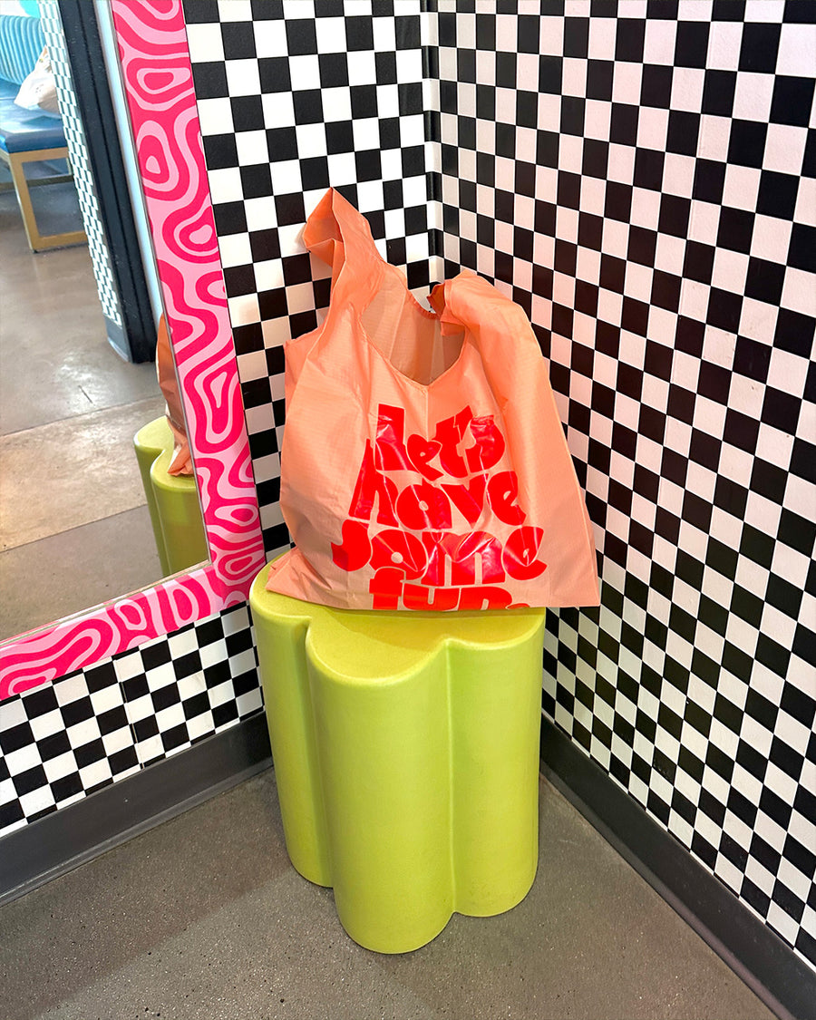 pink standard baggu with red 'lets have some fun' across the front on a flower shaped stool leaning on a black and white checkered wall