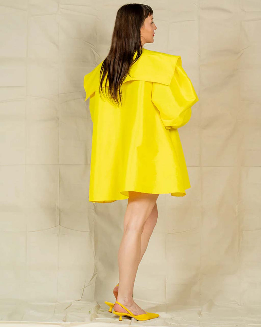 back view of model wearing yellow dress with flowy skirt, oversized sleeves and collar and large yellow bow on the front