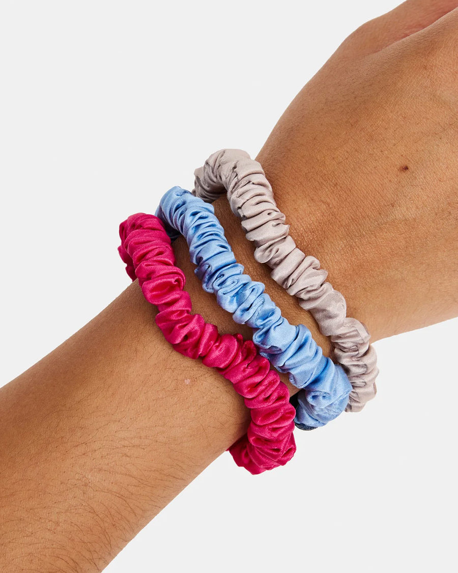 model wearing set of three skinny silk hair scrunchies: grey, hot pink and periwinkle on their wrist