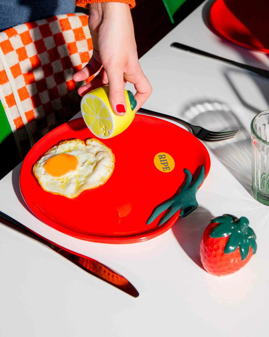 model putting pepper on her egg on a tomato shaped plate with yellow 'ripe' sticker on the left side