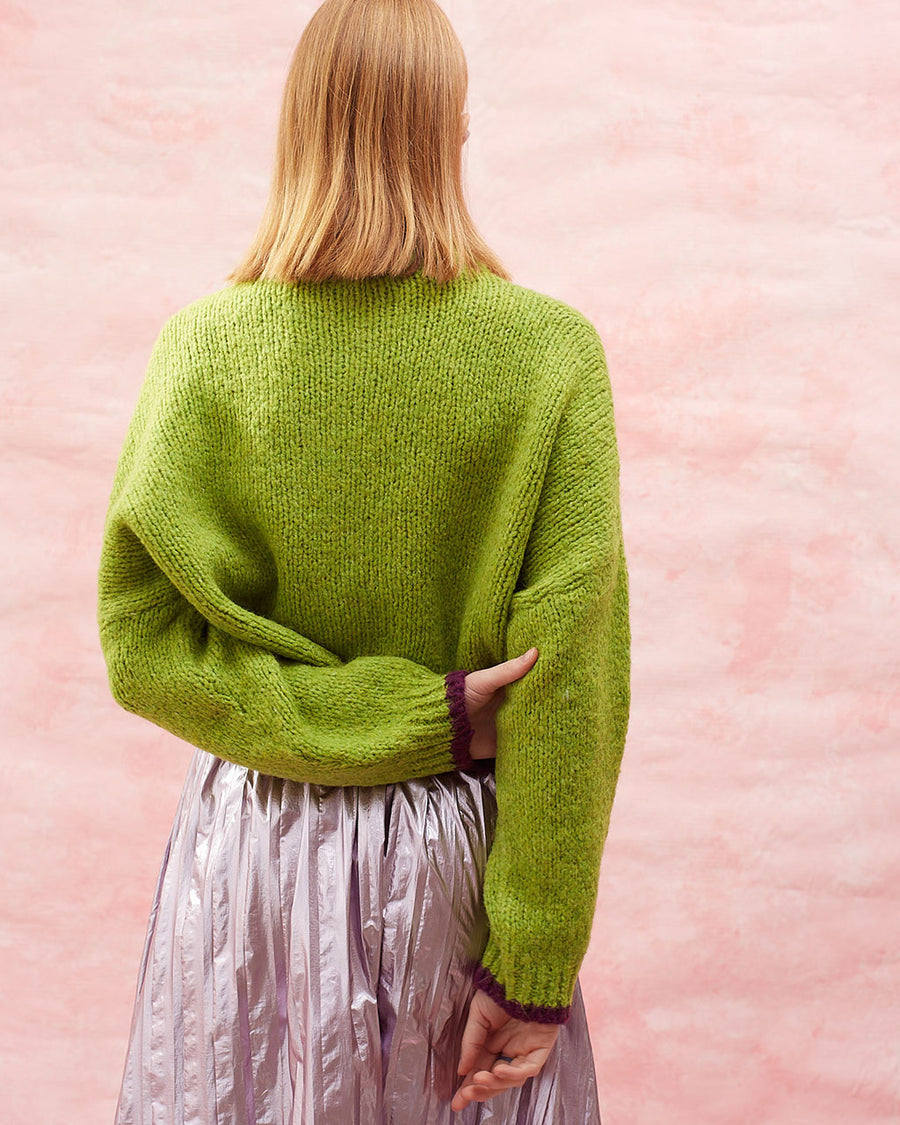 back view of model wearing slightly cropped pea green cardigan with plum patch pockets and trim