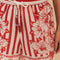 up close of model wearing white and red stripe shorts with beaded tie waist tassels, dolphin hem sides and floral and chain charm print