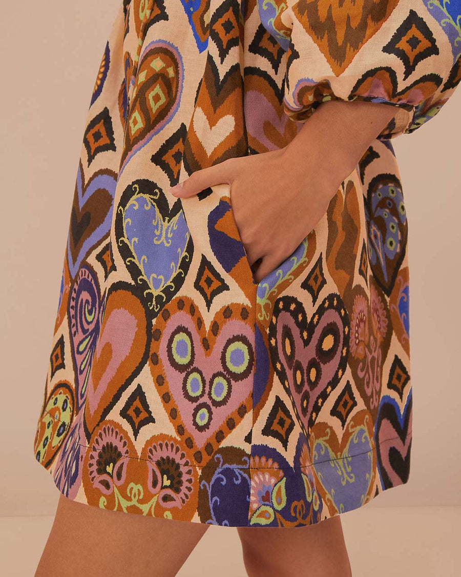 up close of pockets on model wearing square neck mini dress with sand ground and colorful ikat heart print and long puff sleeves