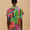 back view of model wearing colorful tropical floral print with button front and pink and green mini tassel detail on the placket and sleeves