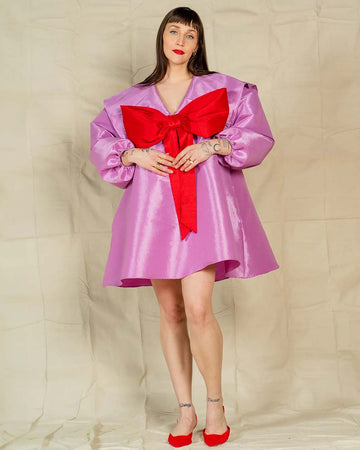 model wearing metallic lavender dress with flowy skirt, oversized sleeves and collar and large red bow on the front