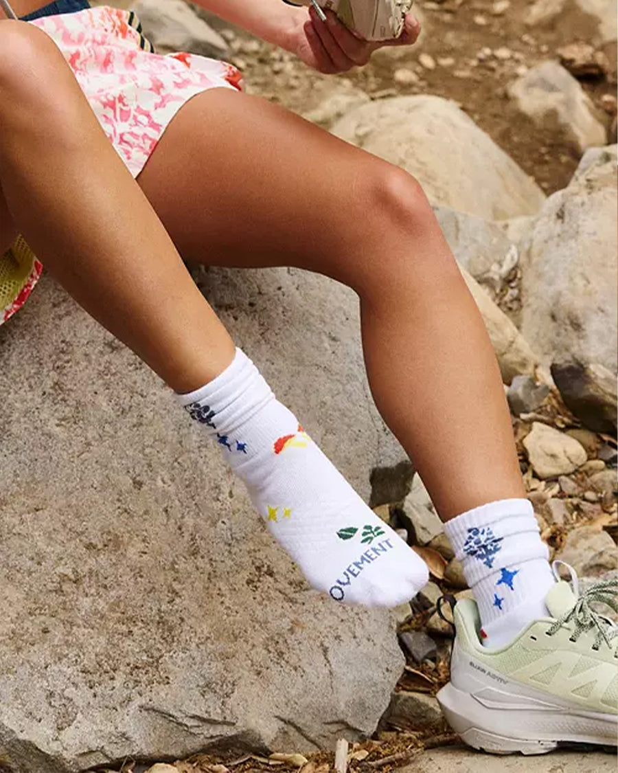 model wearing white long crew socks with colorful leaves, floral and mushroom print sitting on a rock