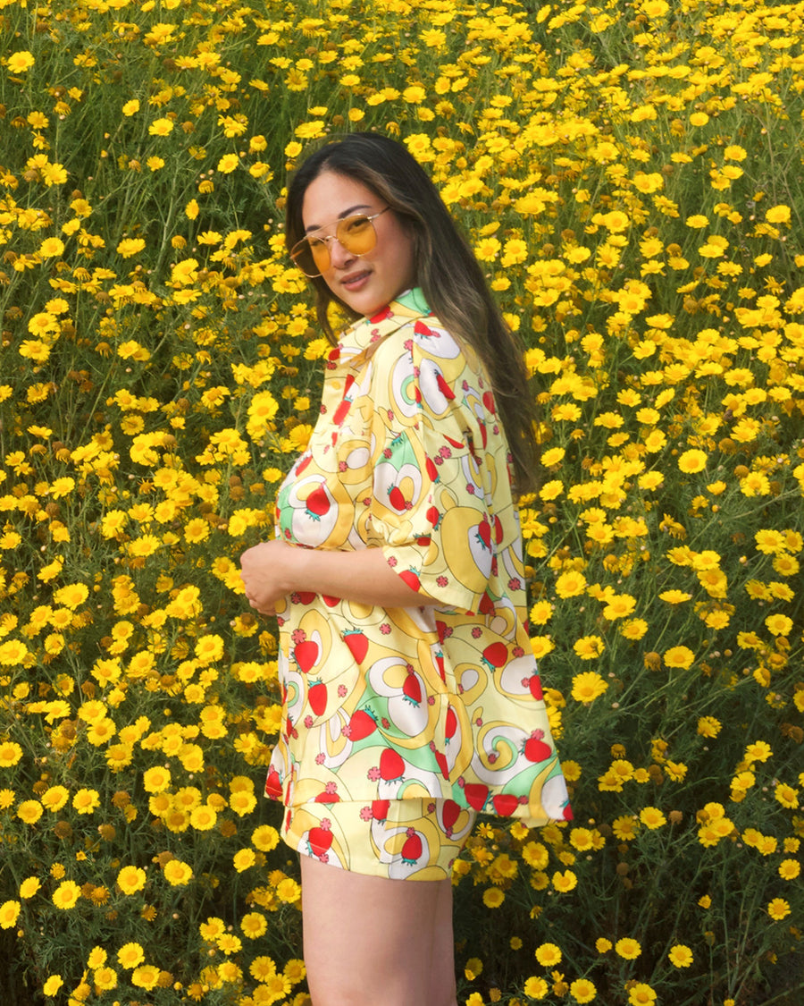 sideview of model wearing yellow trippy button down top with vibrant strawberry print and paired with matching shorts