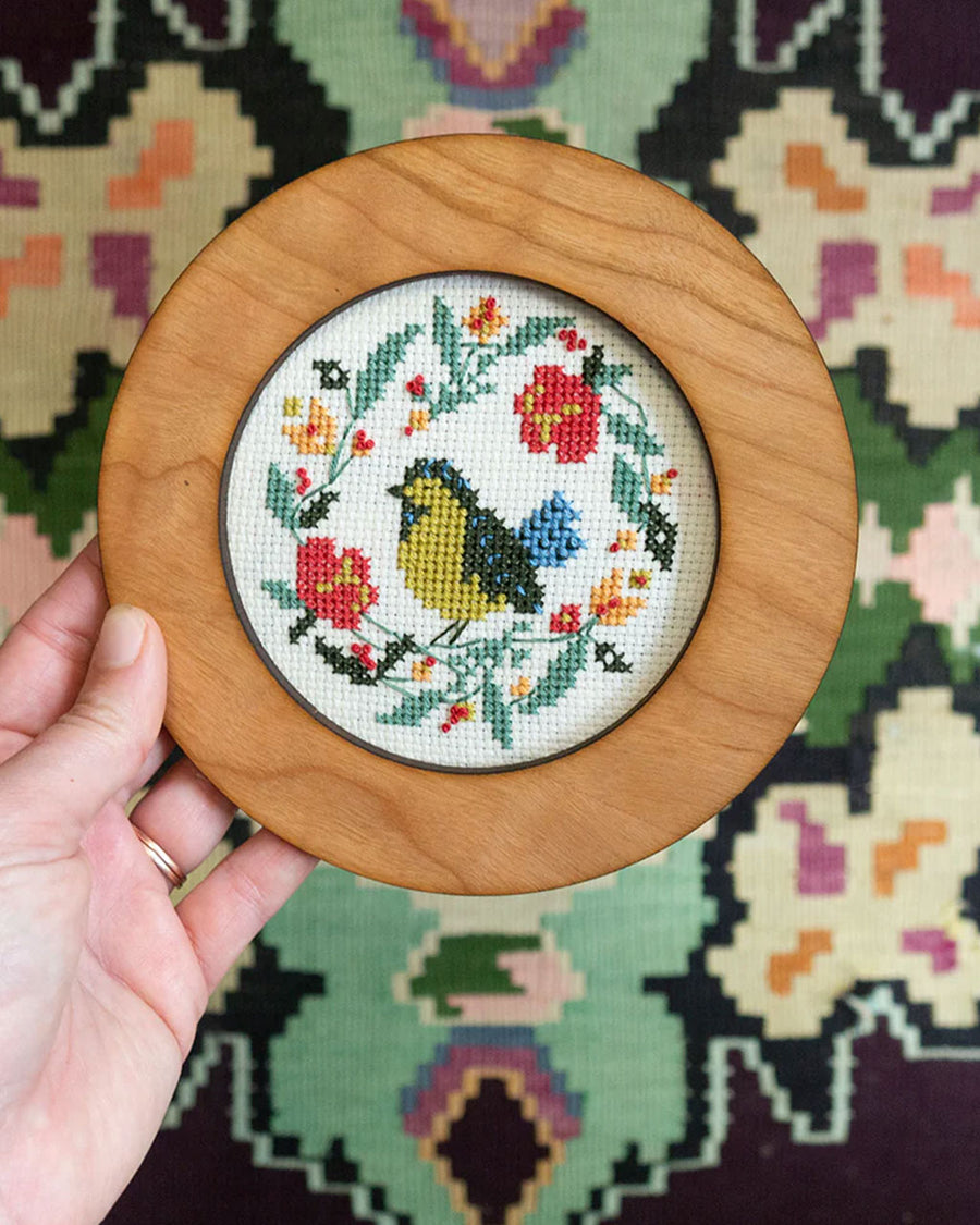 model holding completed model holding white embroidered cross stitch kit with yellow and black bird surrounded  by flowers and has a wood frame