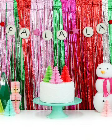 pink red, green and silver metallic fringe backdrop with a tree cake and various holiday decorations on a table