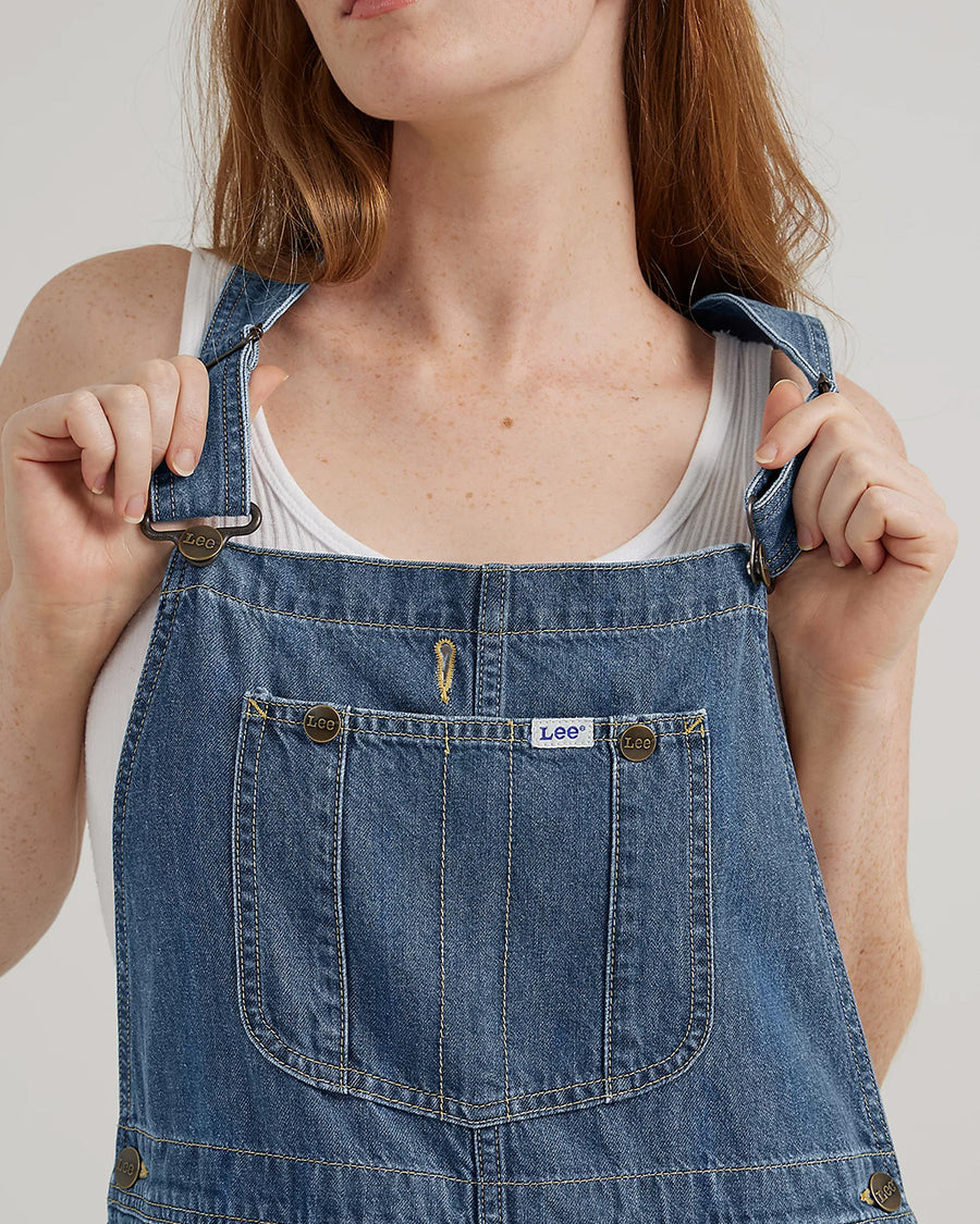 up close of model wearing medium denim overalls with front pocket and lee tag