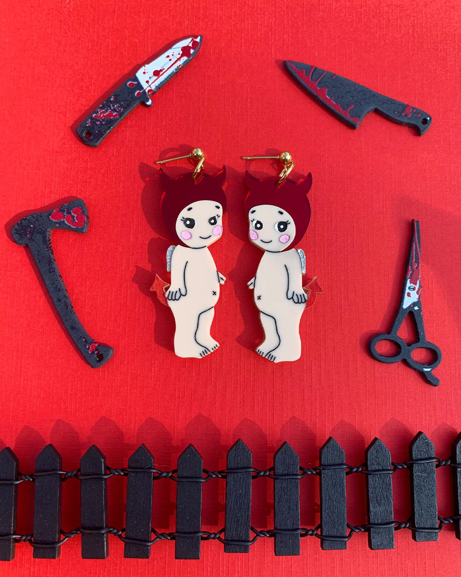 baby wearing devil horn hat and tail dangle earrings surrounded by knives, axe, and scissors