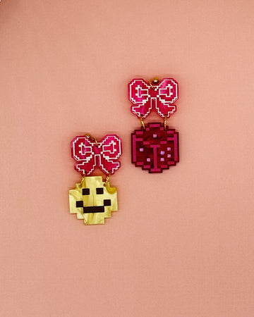 earrings with pink bow stud and one earring with a yellow smiley dangle and a pink dice dangle