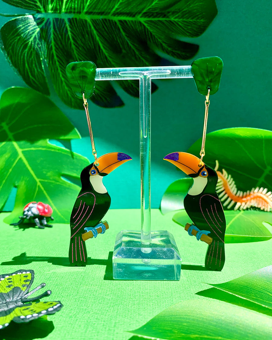 toucan shaped dangle earrings with green leaf stud hanging on a stand