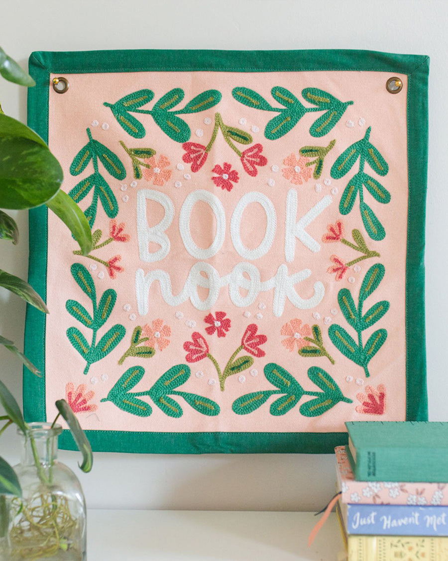 14 in. x 14 in. colorful embroidered wall hanging with 'book nook' across the front displayed on a wall with books