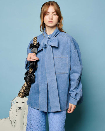 model wearing washed denim long sleeve top with oversized bow on the neckline, front pocket and relaxed fit