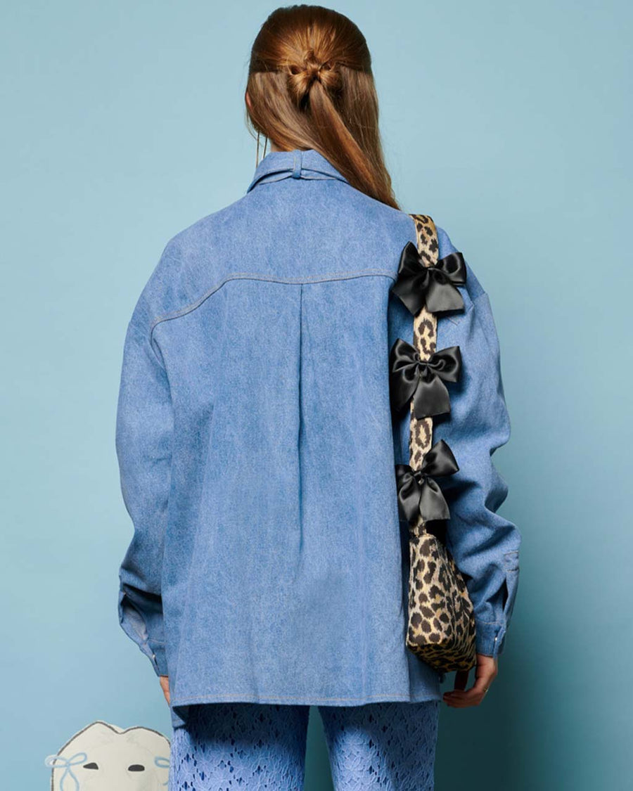 back view of model wearing washed denim long sleeve top with oversized bow on the neckline, front pocket and relaxed fit