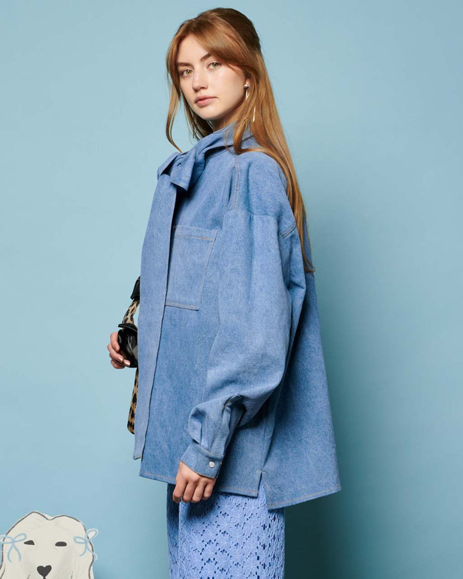 side view of model wearing washed denim long sleeve top with oversized bow on the neckline, front pocket and relaxed fit