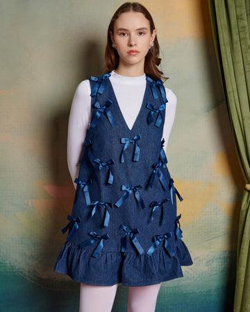 model wearing denim mini dress with ruffle hem skirt, deep v-neckline and all over satin bows