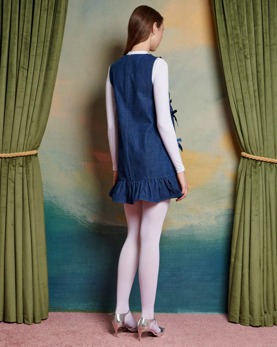 back view of model wearing denim mini dress with ruffle hem skirt, deep v-neckline and all over satin bows