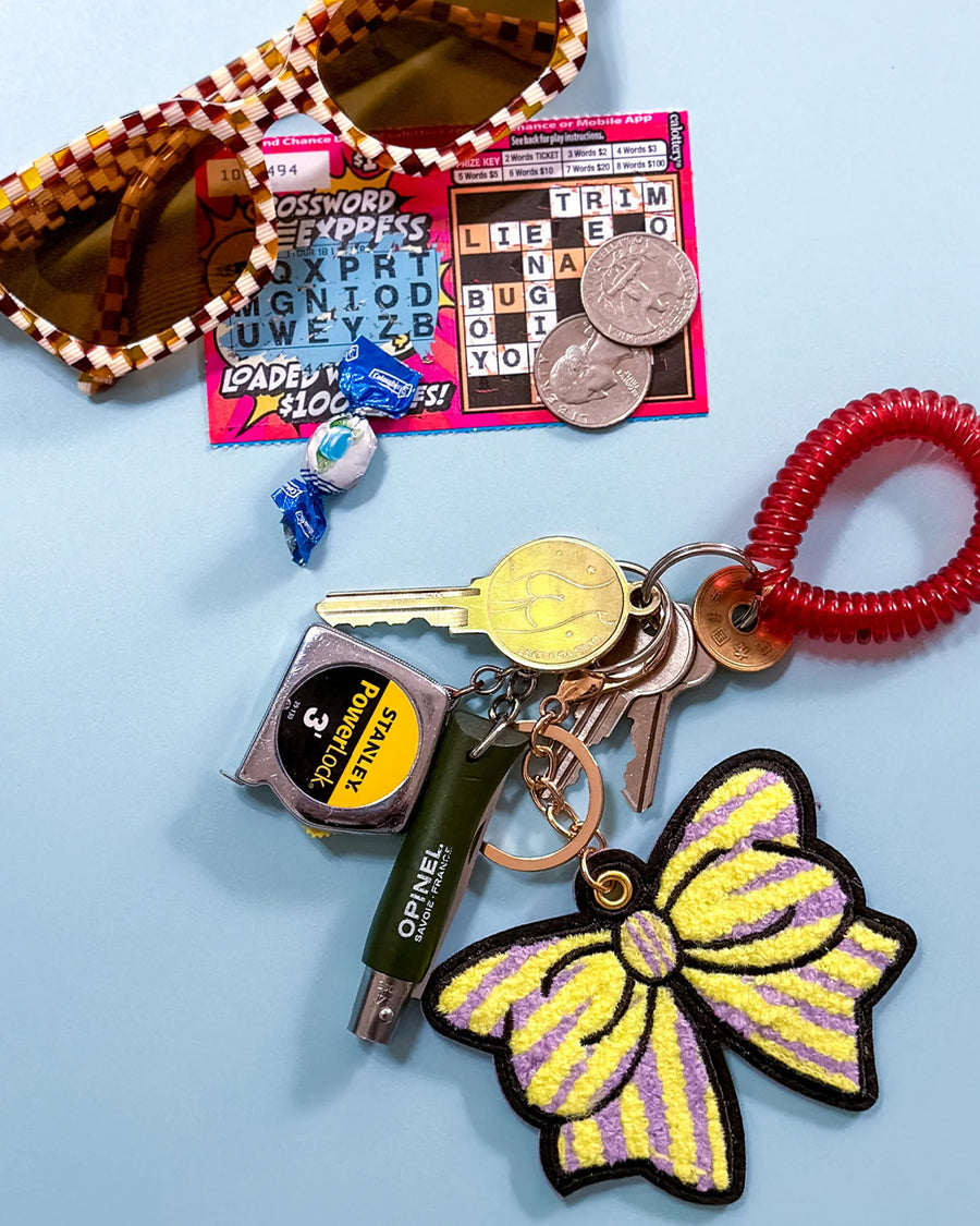 yellow and purple striped keychain with oversized chenille bow on keys with sunglasses and a lotto ticket next to it