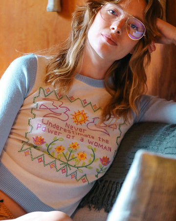 model wearing white sweater with blue sleeves and trim with colorful embroidered 'never underestimate the power of a woman'