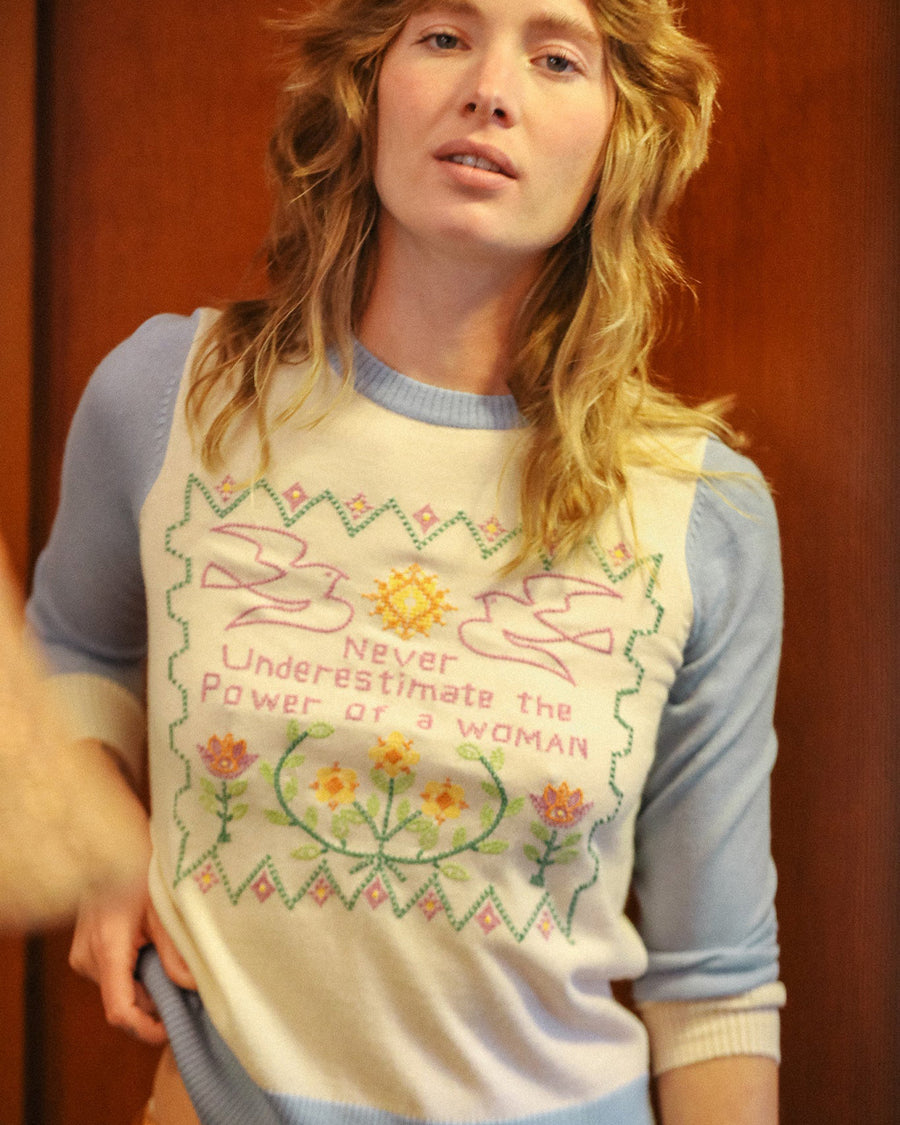 up close of model wearing white sweater with blue sleeves and trim with colorful embroidered 'never underestimate the power of a woman'