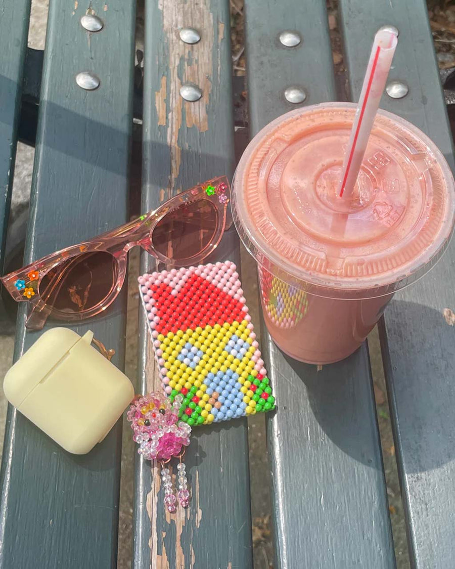 beaded cardholder with yellow house and pink background on a table with a smoothie, sunglasses, and eye buds