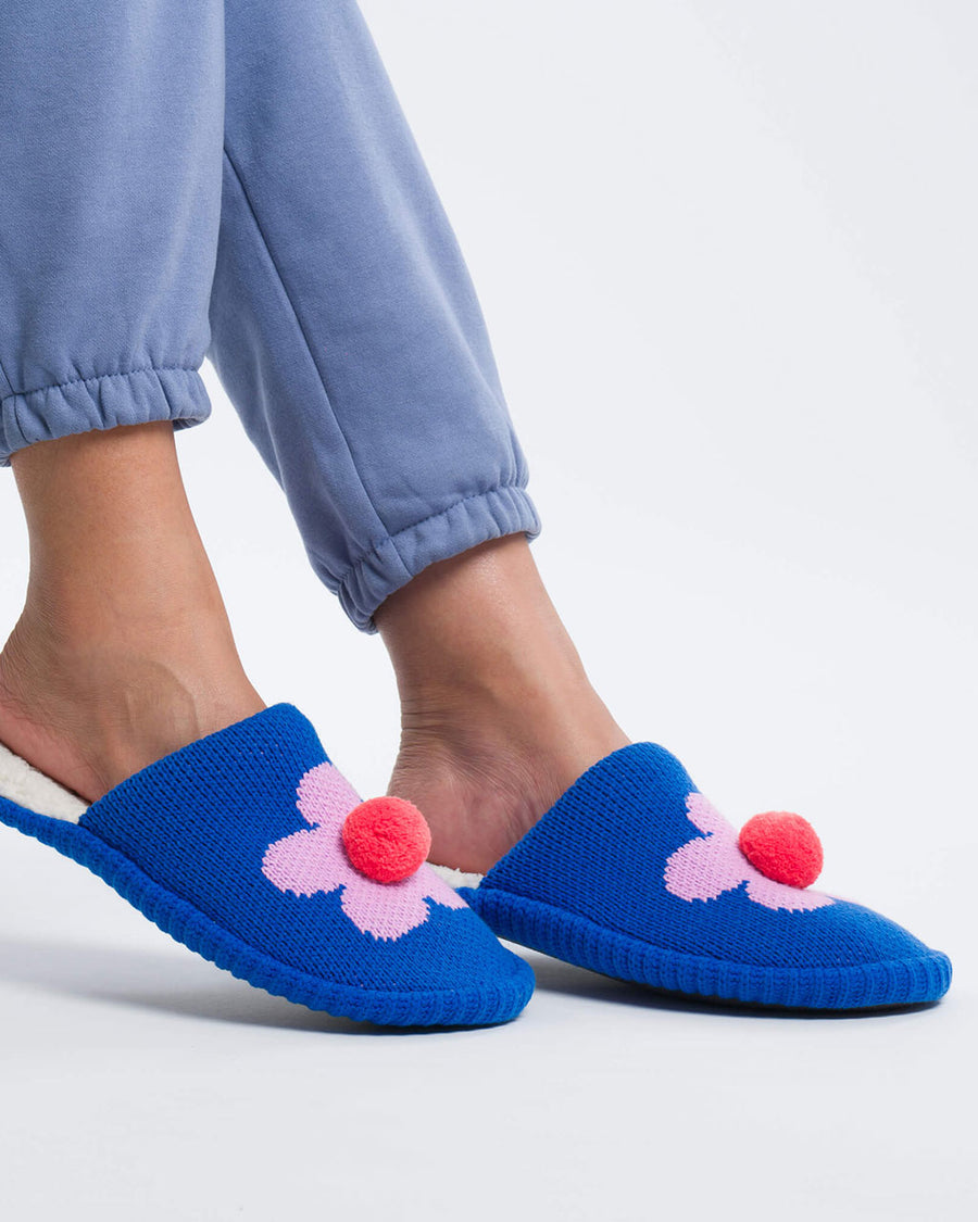 model wearing blue slip on slippers with pink flower and pom detail and fuzzy interior